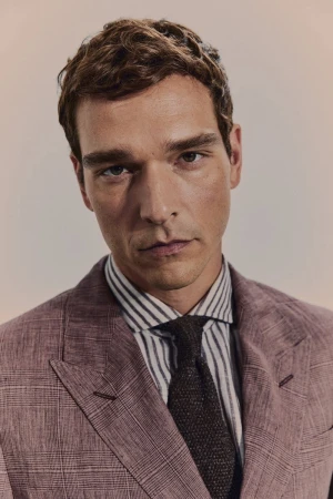 A man in a mauve suit with brown hair gazes forward, his striped shirt and tie complementing the formal yet approachable style.