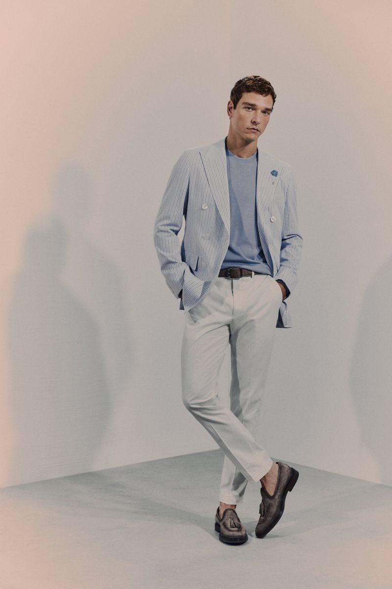 A stylish adult male model in a striped blazer, light blue t-shirt, and white pants poses in a corner, showcasing high-end fashion.