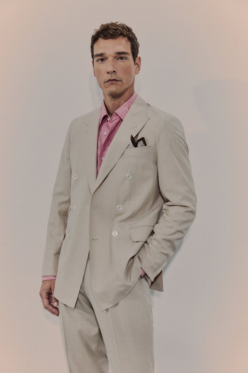 An adult male poses indoors, wearing a beige suit and a pink patterned shirt. The shot is a medium, front-facing view against a neutral background.