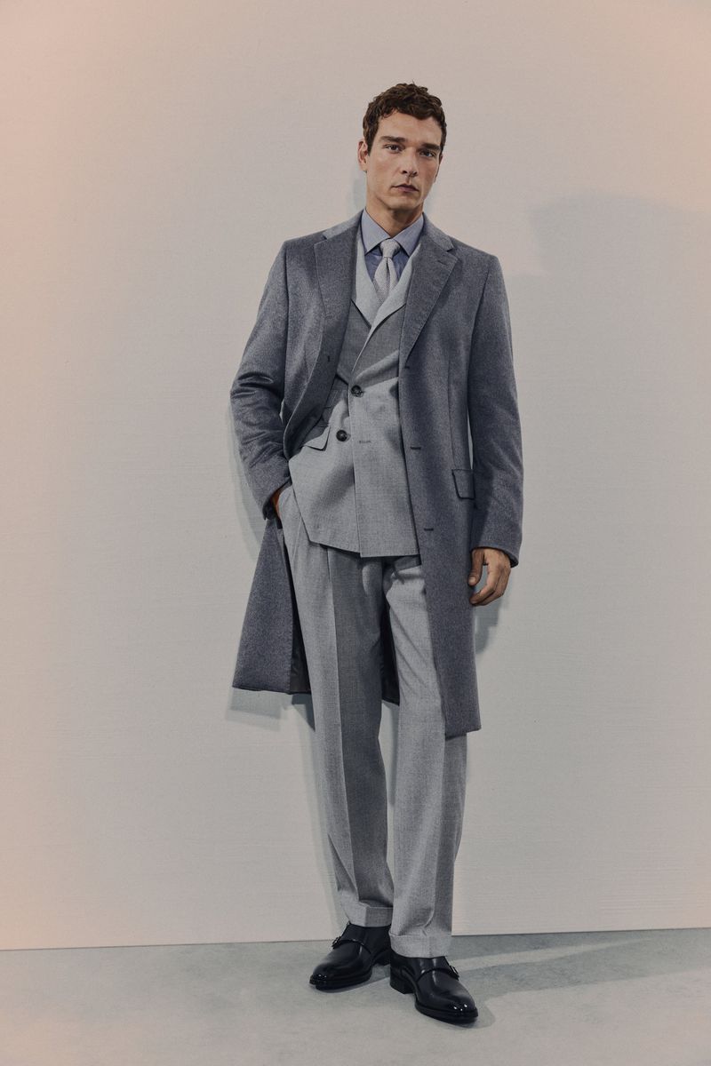 Full-length portrait of a male fashion model in a gray three-piece suit, overcoat, dress shirt, and black leather shoes posing indoors.