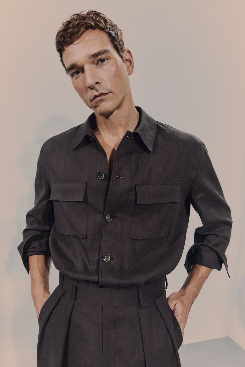 Male model posing in a dark-grey, button-down shirt with patch pockets and matching tailored pants on a neutral backdrop, studio shot.