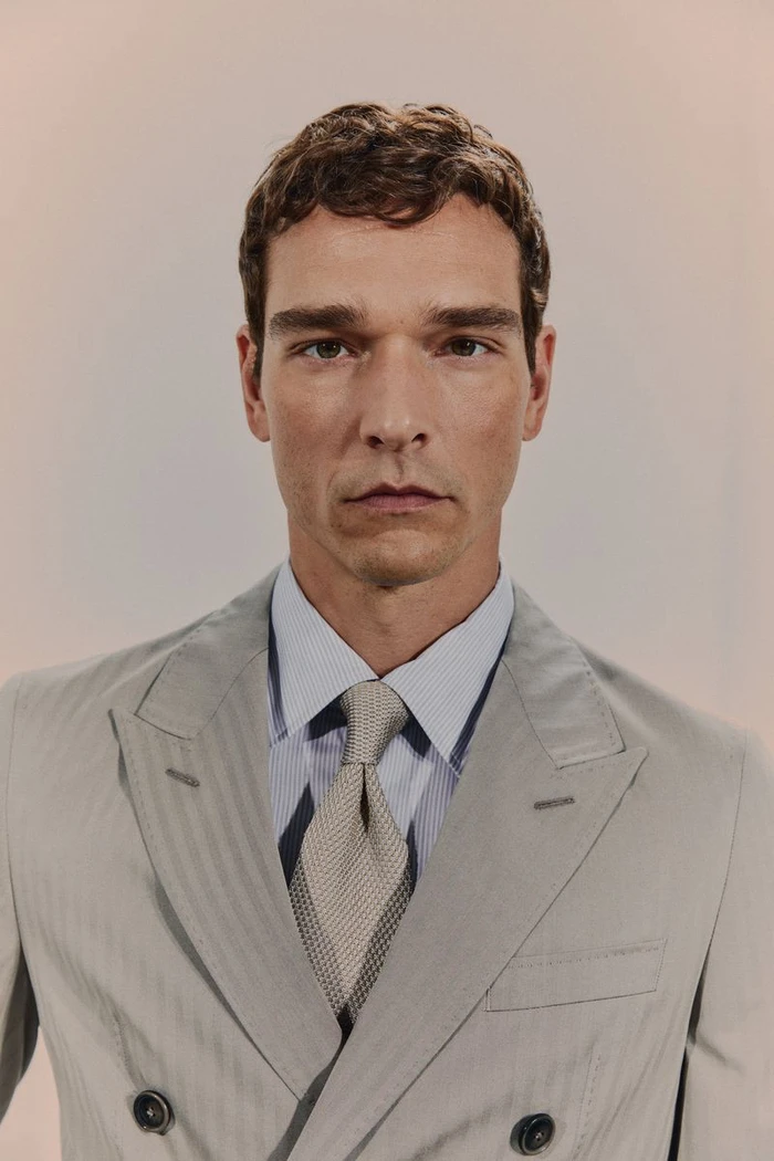 Portrait of a man wearing a light grey double-breasted suit with a striped shirt and textured tie against a neutral background.