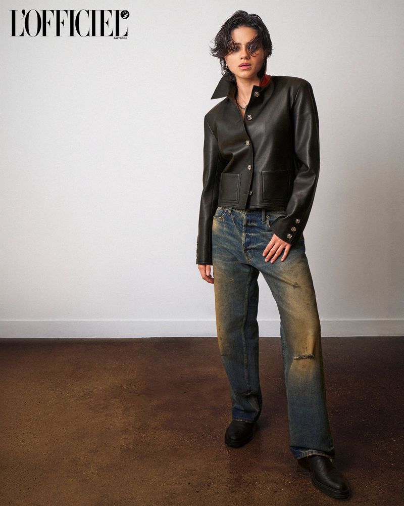 Sasha Calle standing against a white studio wall wearing a black leather jacket, distressed blue jeans, and black shoes.