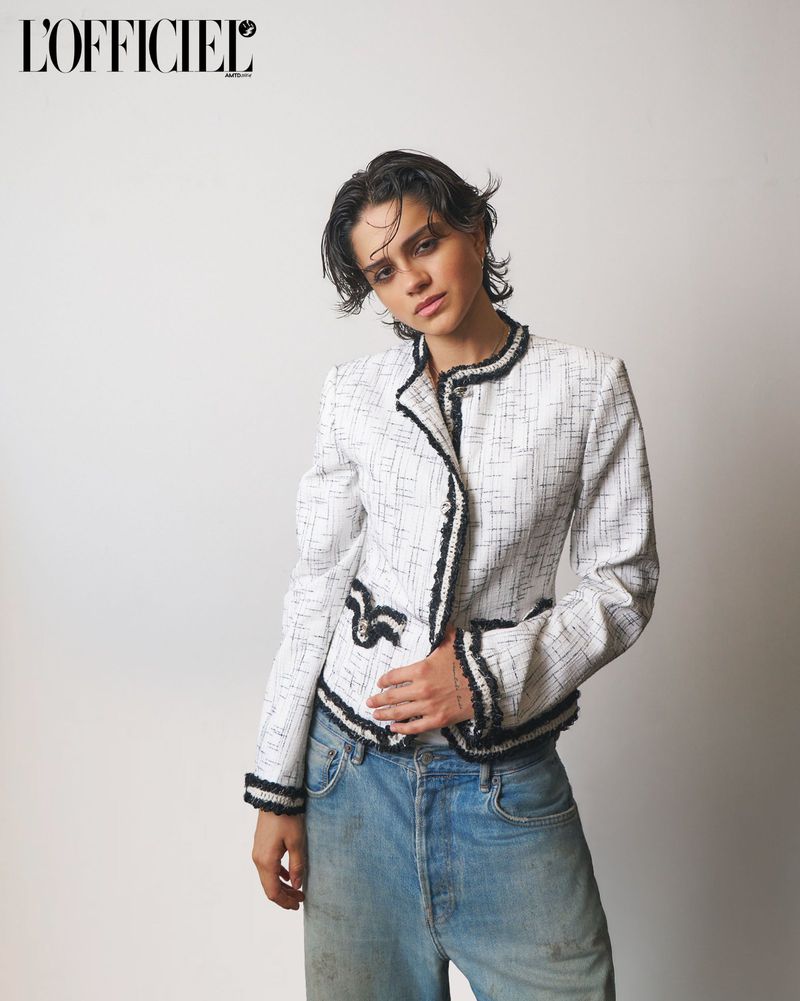 Actress Sasha Calle posing in a white textured tweed jacket with black trim and relaxed blue jeans against a studio backdrop.