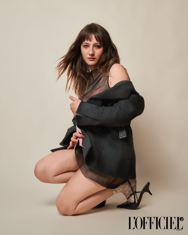 Actress Lily Collias kneeling in a dark tailored jacket and sheer layered outfit against a warm studio backdrop for L'Officiel USA.