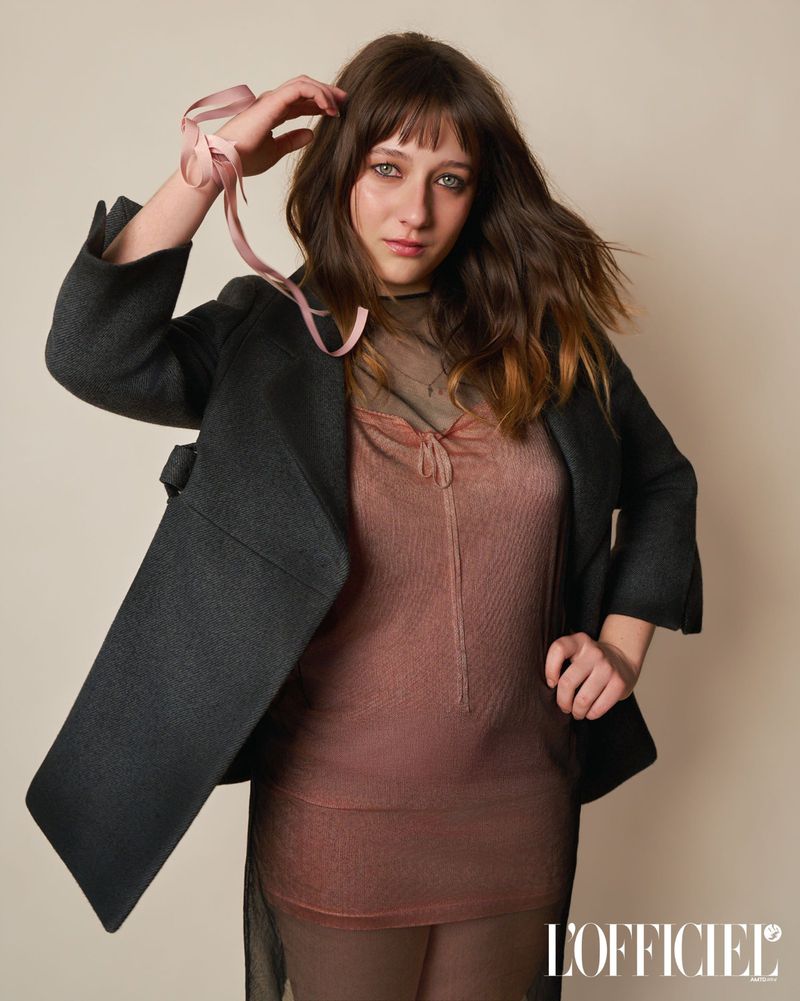 Lily Collias posing in a charcoal blazer and sheer dusty rose tunic dress with a pink ribbon on her wrist against a studio backdrop.