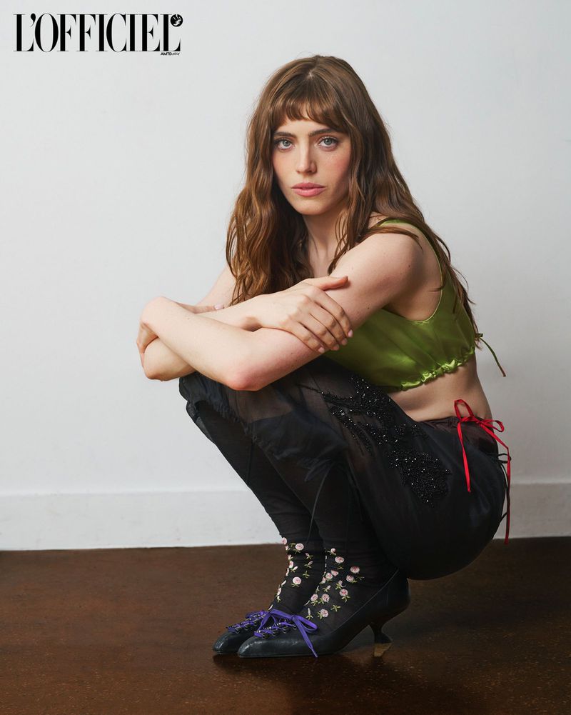 Bella Maclean crouching in a green crop top, embellished black trousers, and pointed heels against a white wall.