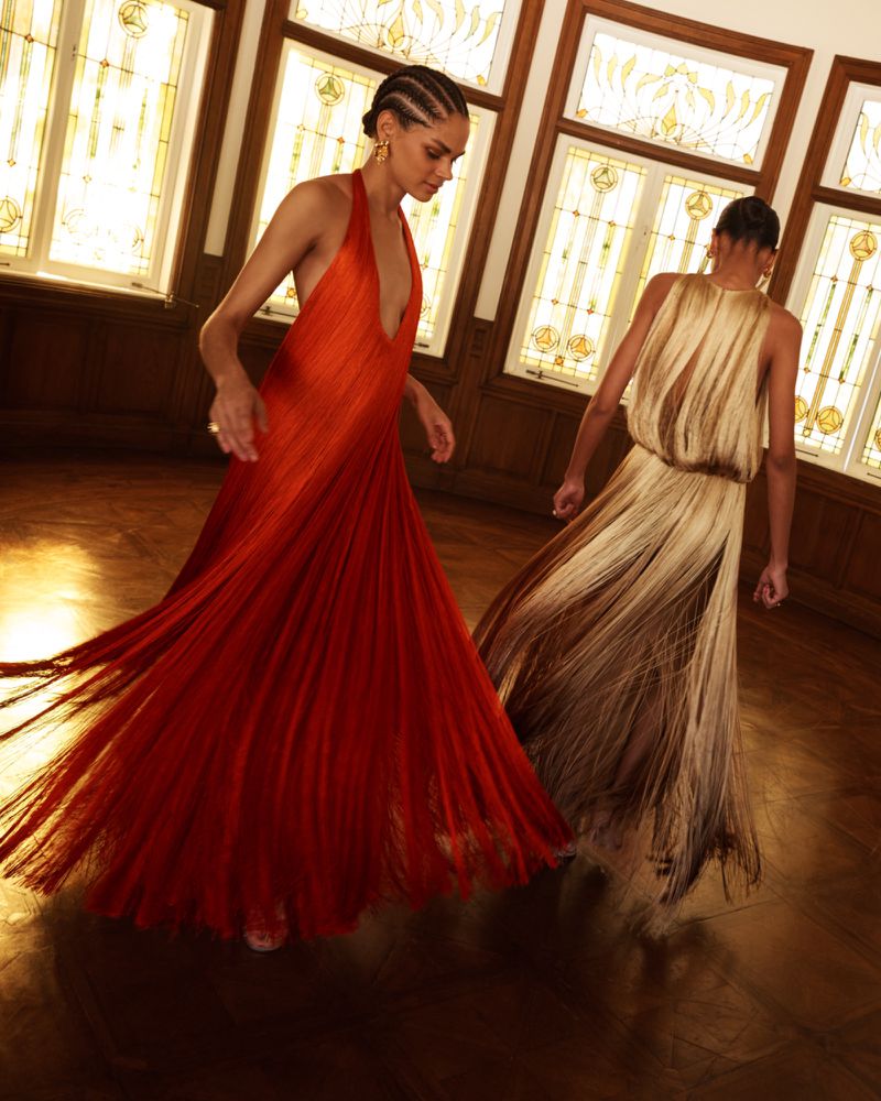 Two models wearing flowing fringed dresses in an interior room with large stained glass windows