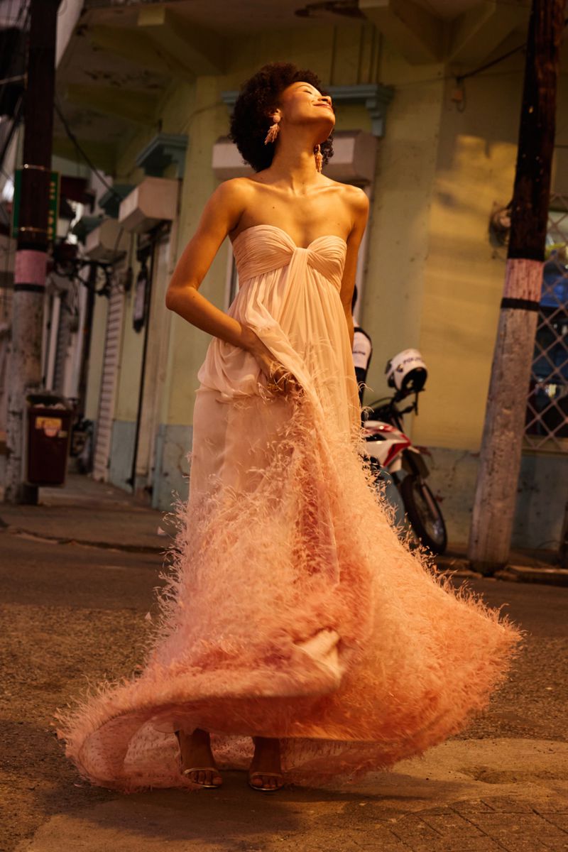 Model Lineisy Montero twirls in a strapless peach feather-trimmed evening gown on an urban street at dusk.
