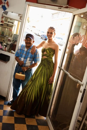 Model in a strapless green gown standing beside an older man holding a gold clutch inside a barbershop