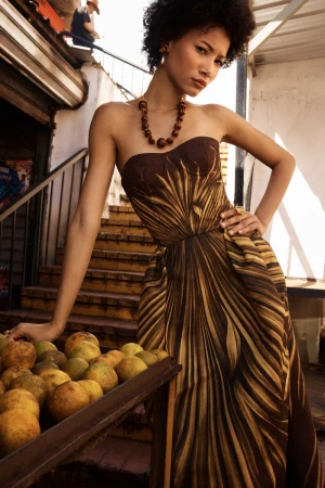 Model posing in a strapless brown palm-print gown beside a fruit cart on outdoor steps.