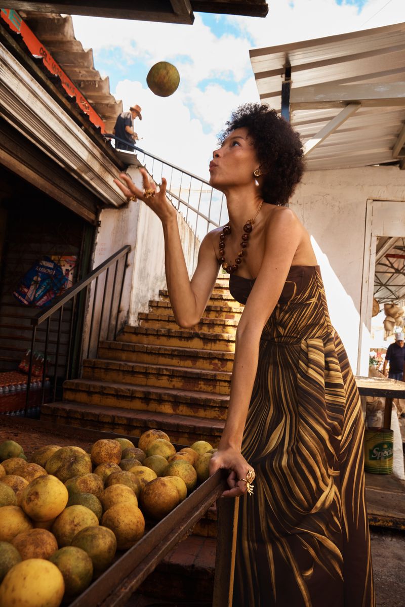Model in brown patterned strapless dress tossing a round fruit near a market stall with citrus fruit crates.
