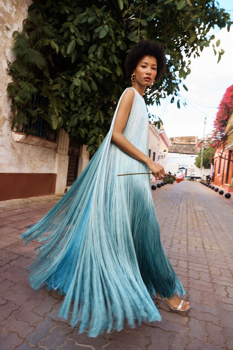 Model in a flowing blue gradient dress walking on a cobblestone street lined with buildings and lush foliage