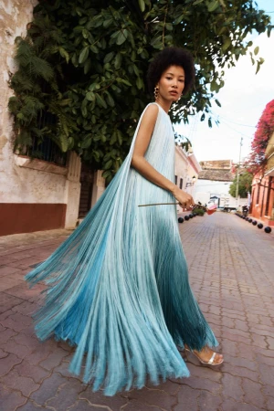 Model in a flowing blue gradient dress walking on a cobblestone street lined with buildings and lush foliage