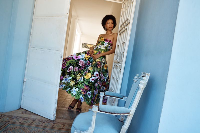 Model wearing a strapless floral print midi dress leaning against a white doorway beside a light blue armchair on a tiled floor.