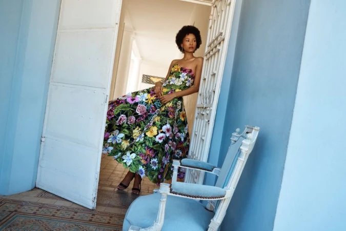 Model wearing a strapless floral print midi dress leaning against a white doorway beside a light blue armchair on a tiled floor.