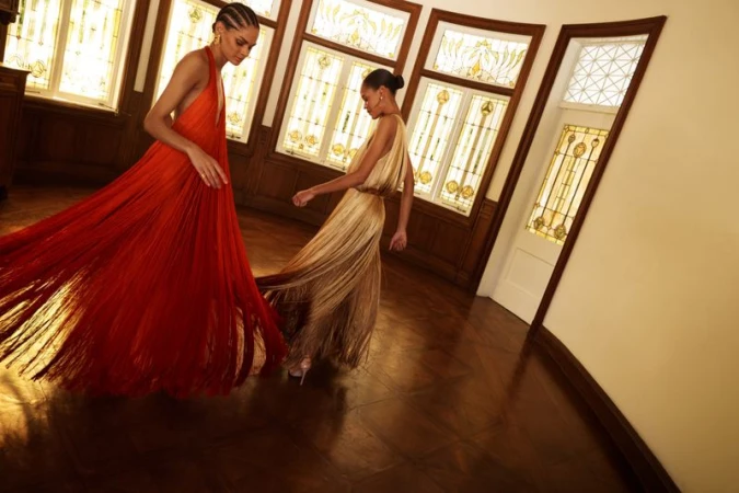 Two fashion models spinning in long red and gold fringe gowns across a polished wood parquet floor with stained-glass windows.