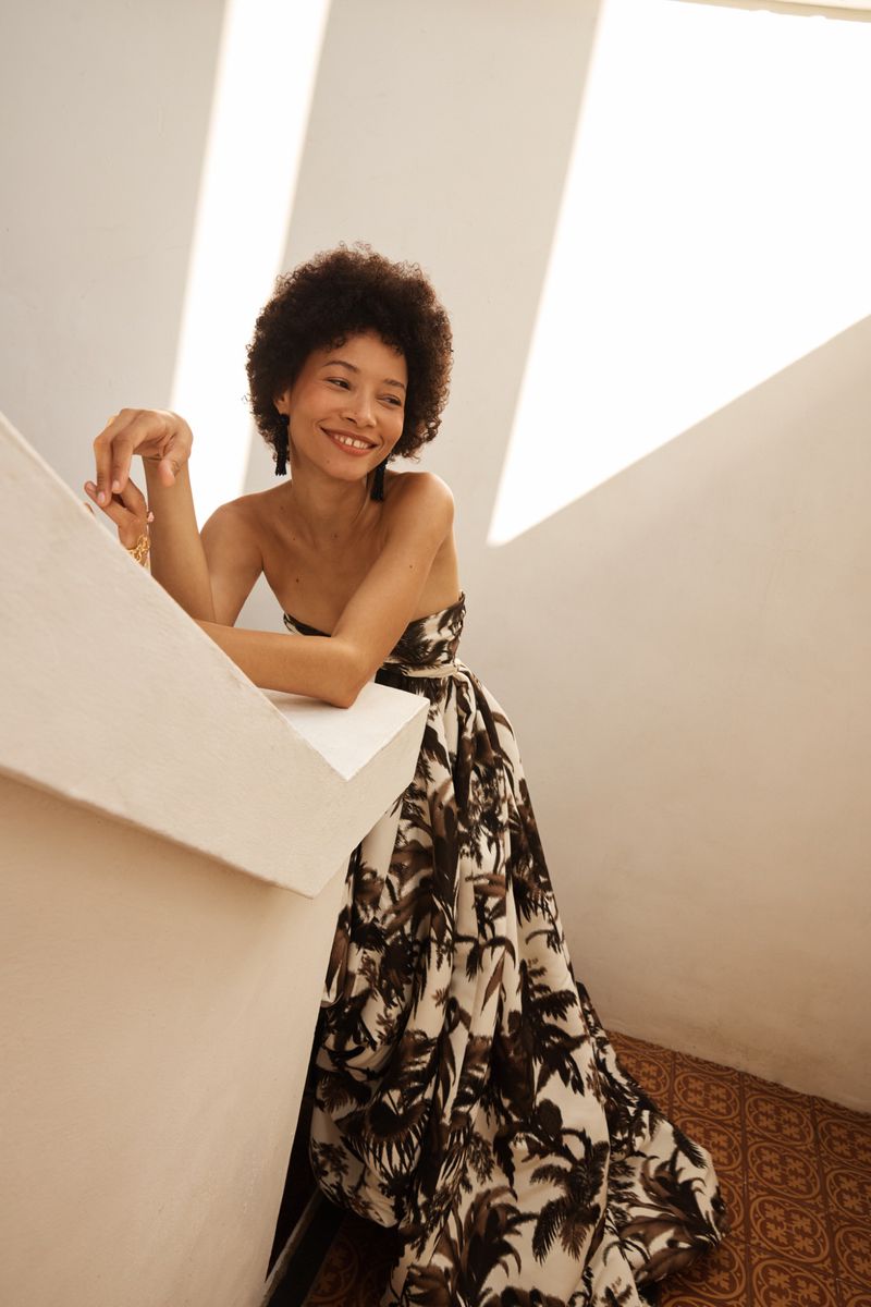Model smiling in a strapless palm tree print ballgown while leaning against a white architectural wall in bright sunlight.