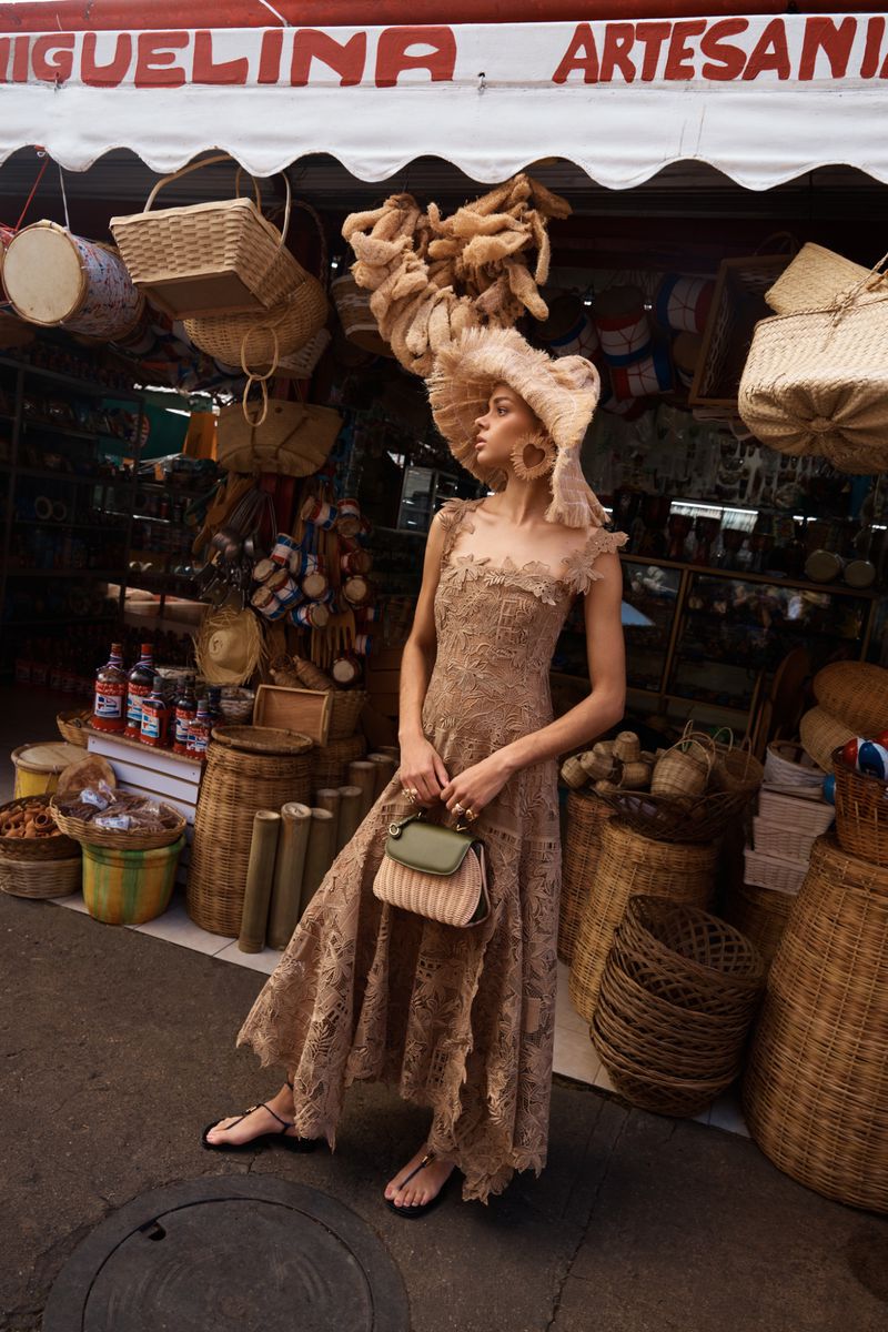 Model wearing a tan lace dress and wide-brim frayed raffia hat holding a handbag in front of a market stall.