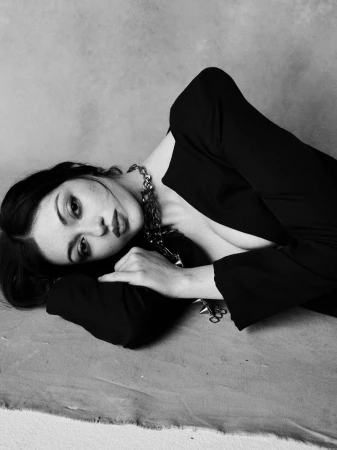 Black and white portrait of a woman reclining in a black tailored blazer and chunky metal chain necklace on a canvas backdrop.