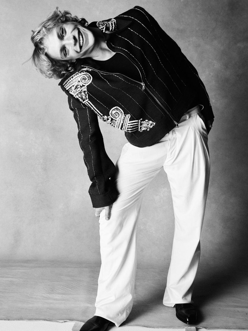 Black and white portrait of a person in an embroidered dark jacket and white trousers posing against a grey seamless studio backdrop.