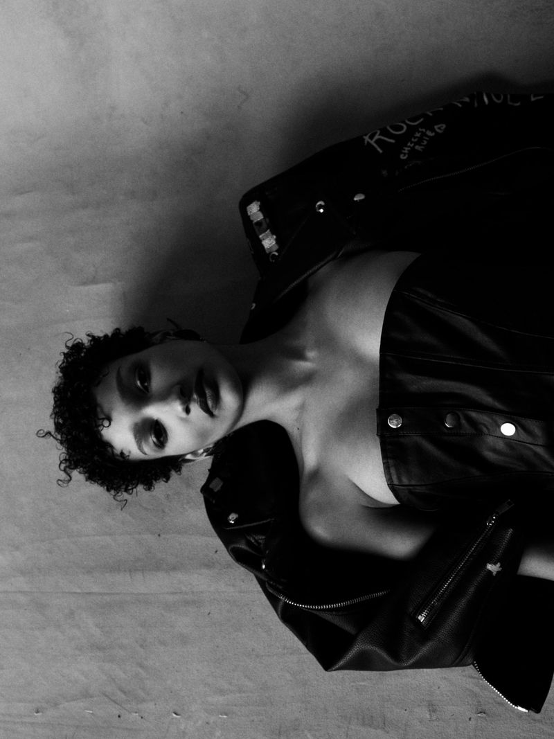 Black and white portrait of model Jules Santiago wearing a black leather jacket and strapless top against a textured studio background.