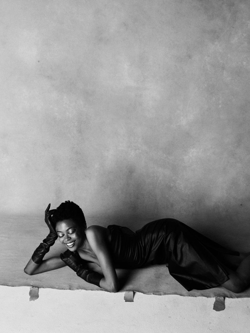 Model lying down in a strapless black leather dress and long leather gloves against a textured studio backdrop.