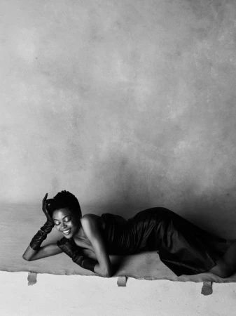 Model lying down in a strapless black leather dress and long leather gloves against a textured studio backdrop.