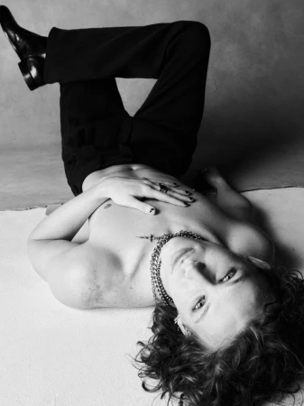 Black and white portrait of a person lying on the floor with legs crossed wearing dark trousers and a chain necklace.