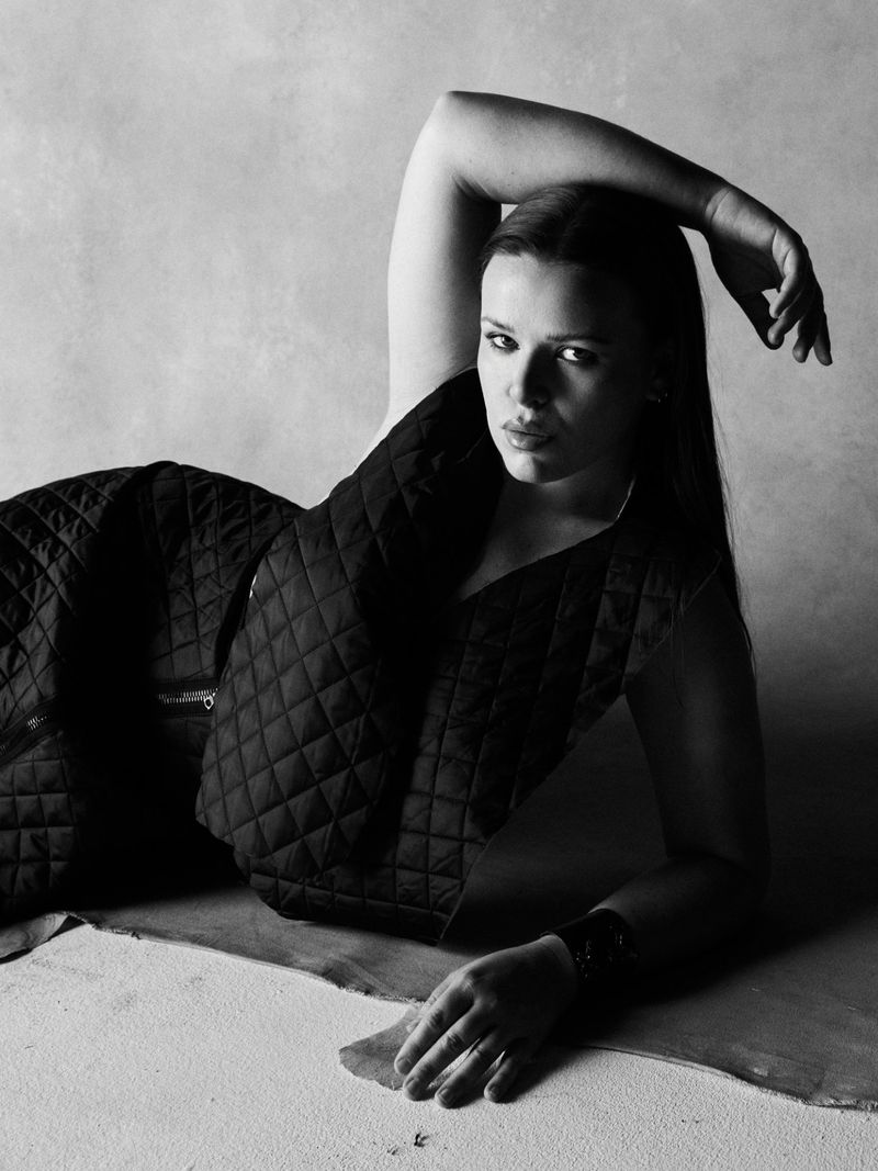 Monochrome fashion portrait of a woman in a black quilted vest reclining on a textured studio floor.