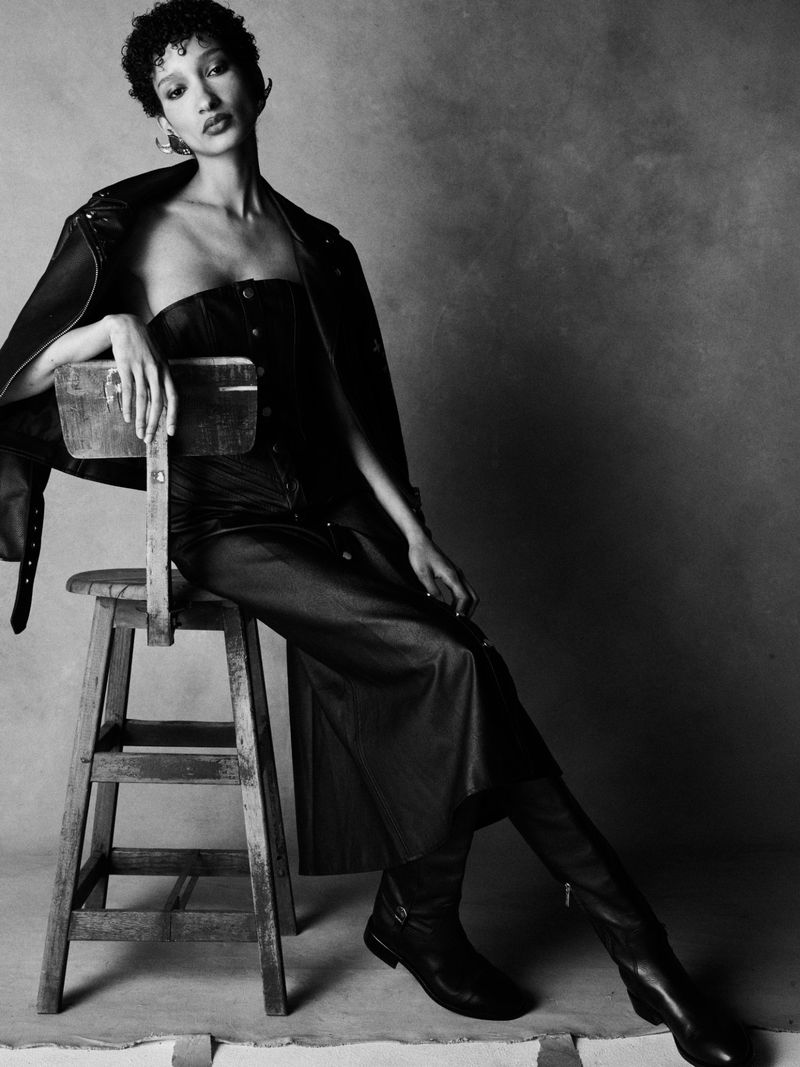 Model seated on a wooden stool wearing a leather jacket and dress against a textured dark background