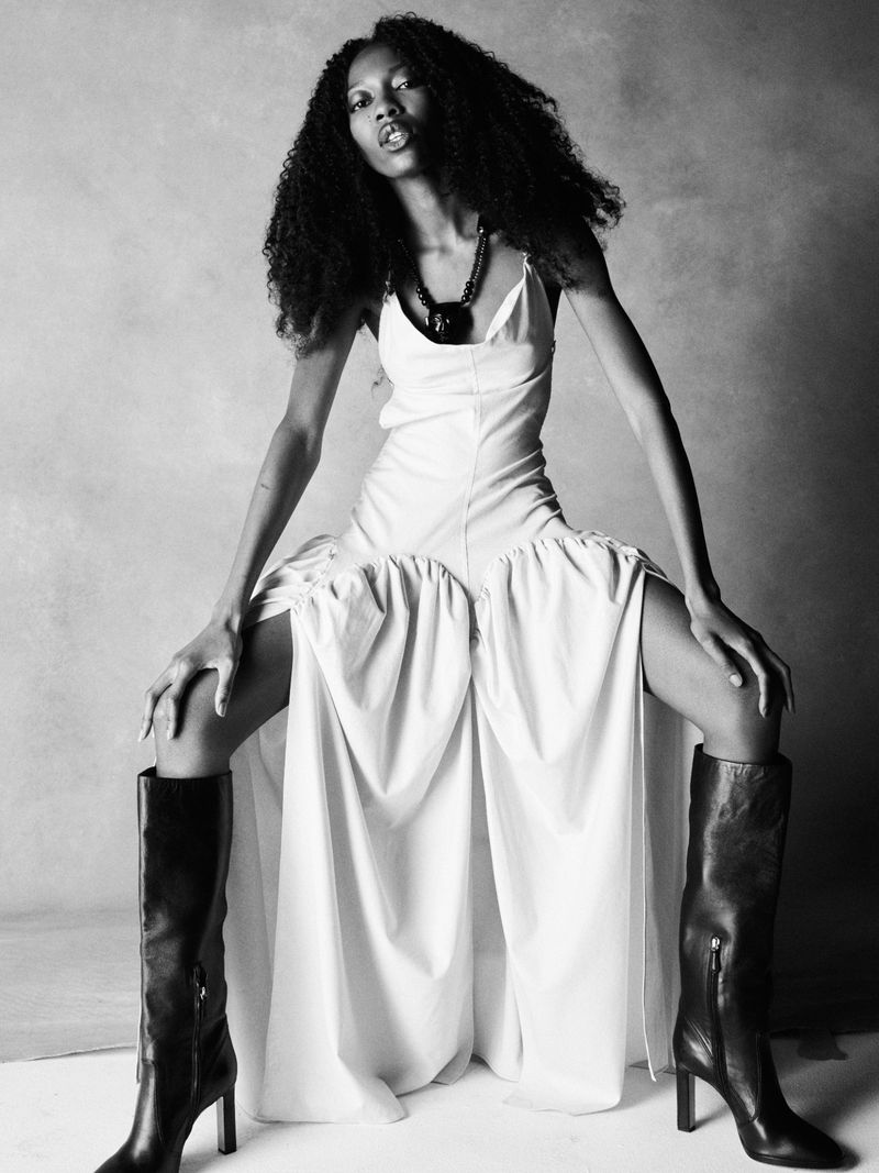 Model Jules Santiago posing in a draped white dress and knee-high black boots against a studio backdrop in black and white.