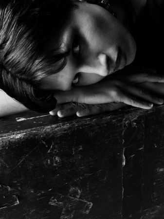 Close-up black and white portrait of a person resting their head and hands on a textured dark surface looking at the camera.