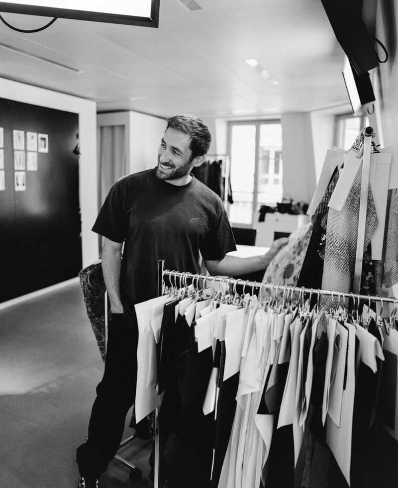 Black and white portrait of designer Antonin Tron smiling beside a garment rack in a fashion studio.