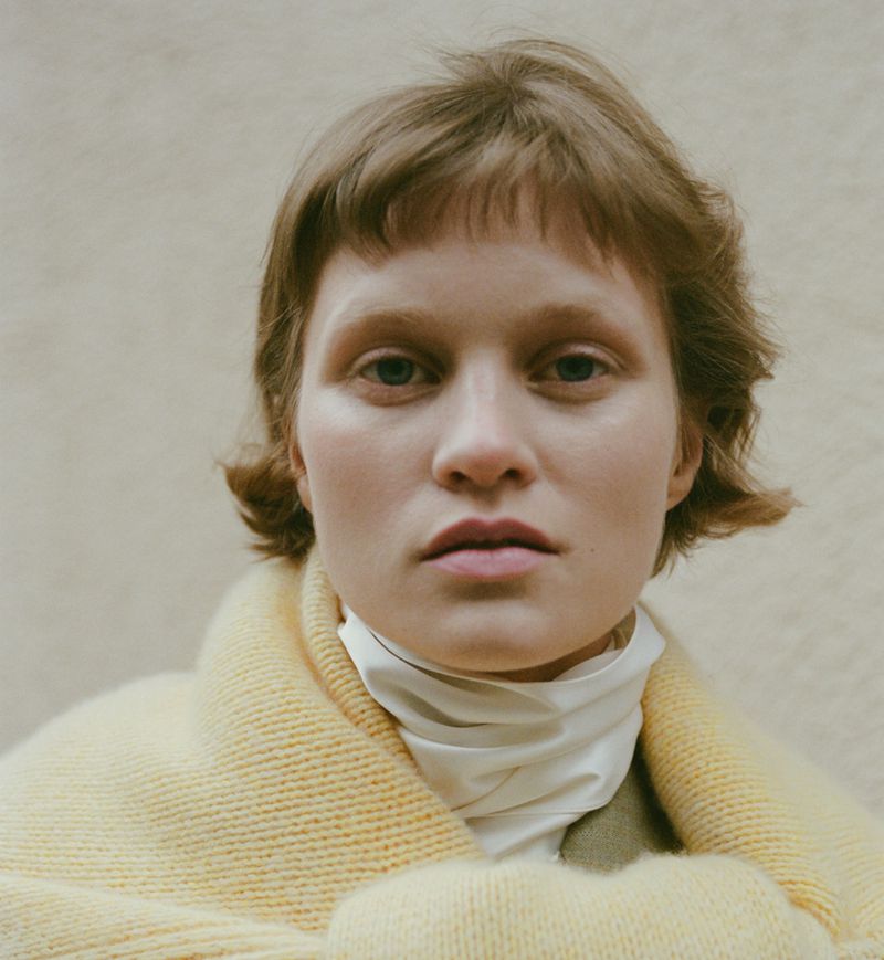 Close-up portrait of model Sofia Hansson wearing a yellow knit shawl and white high-neck collar against a neutral background.