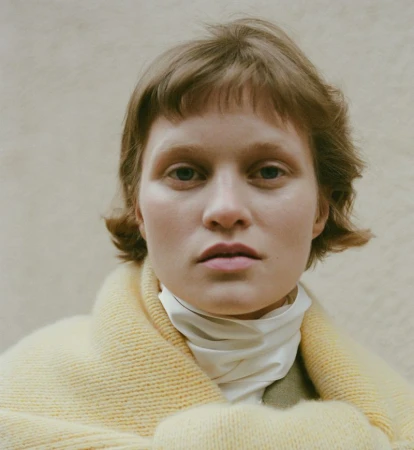 Close-up portrait of model Sofia Hansson wearing a yellow knit shawl and white high-neck collar against a neutral background.