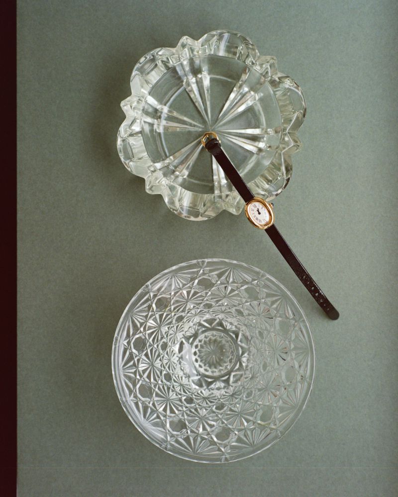 Overhead view of two cut crystal glass dishes and a slender wristwatch with a dark strap arranged on a sage green surface.