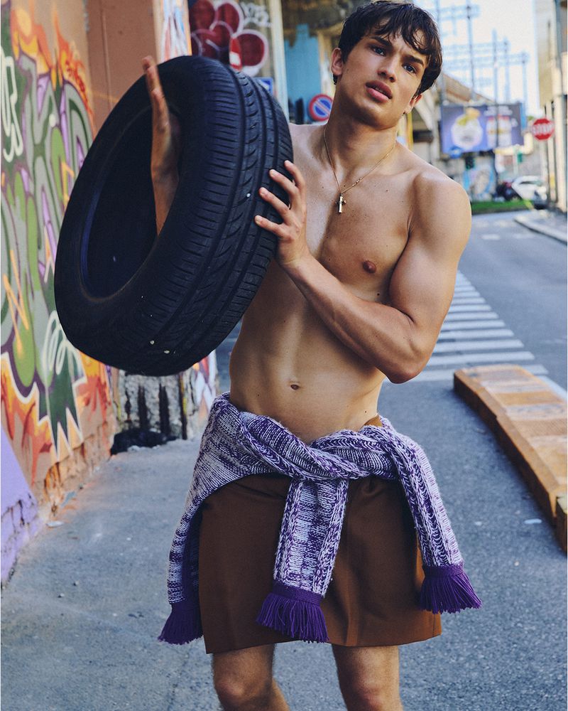 Shirtless male model holding a large car tire against an urban street background with graffiti