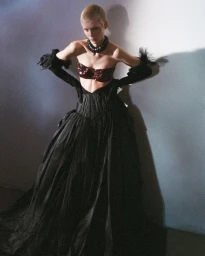 Model Sofia Mathiassen poses in a voluminous black gown, sequined burgundy bra, and black opera gloves against a studio wall.