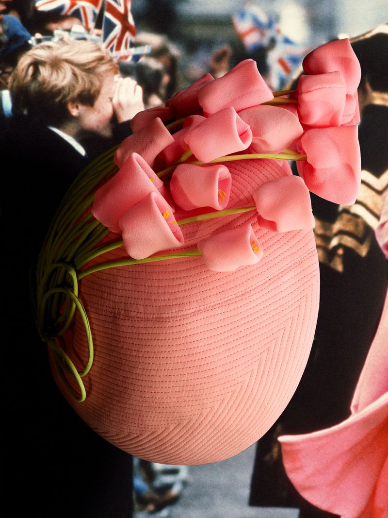 Close-up of a pink textured millinery hat adorned with pink fabric flowers and green stems against a crowd backdrop.