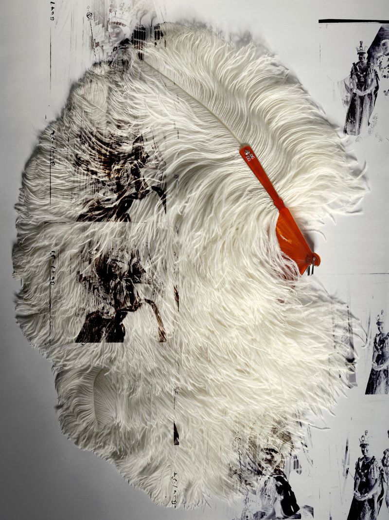 White ostrich feather fan with an orange handle resting on archival monochrome photographs of Queen Elizabeth II.