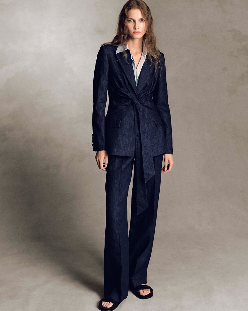 Model wearing a tailored dark navy tie-waist suit and slide sandals against a mottled grey studio backdrop.