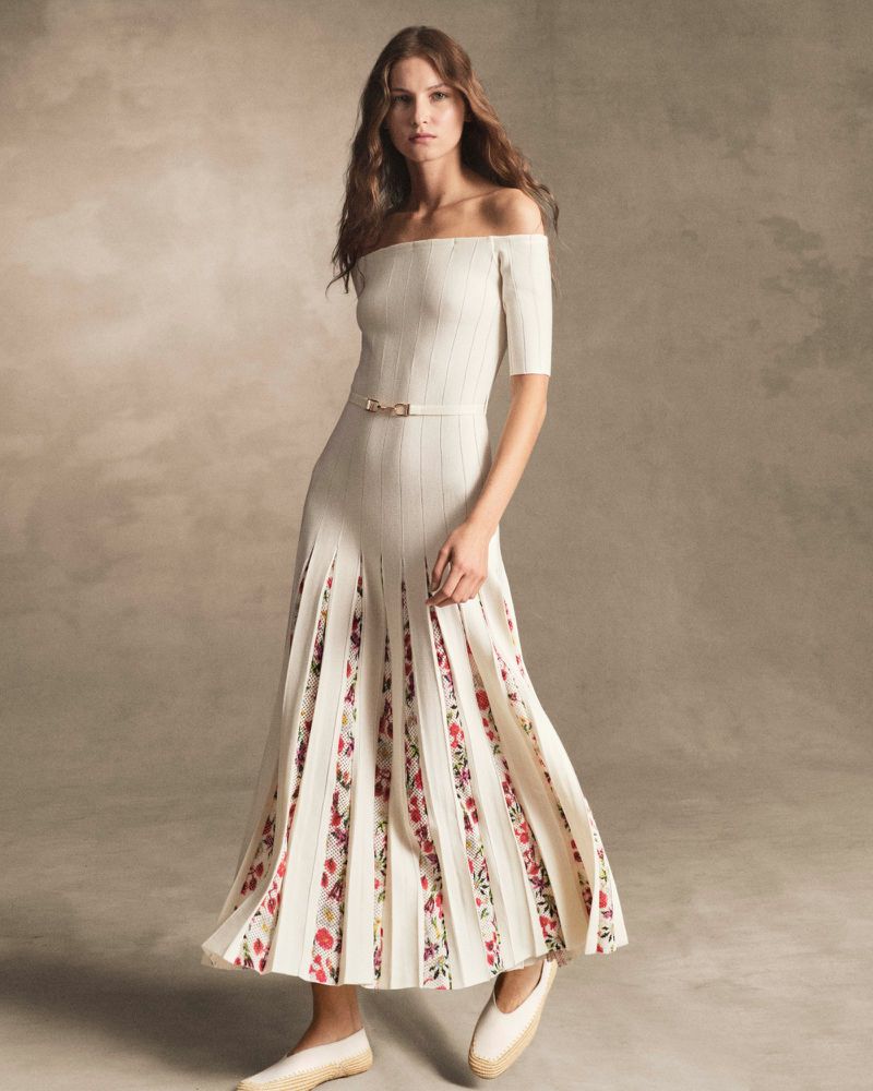 Model wearing an off-the-shoulder cream knit dress with floral pleated inserts against a warm textured studio backdrop.