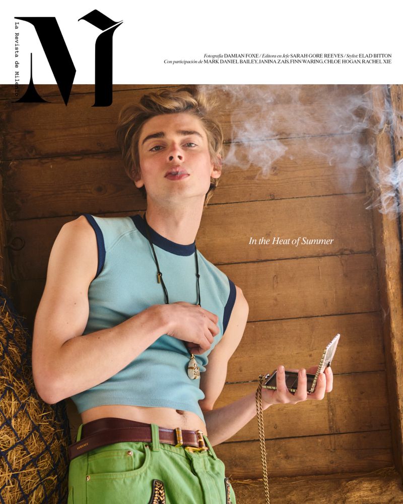 Young male model wearing a light blue sleeveless top and green trousers standing inside a rustic wooden structure.