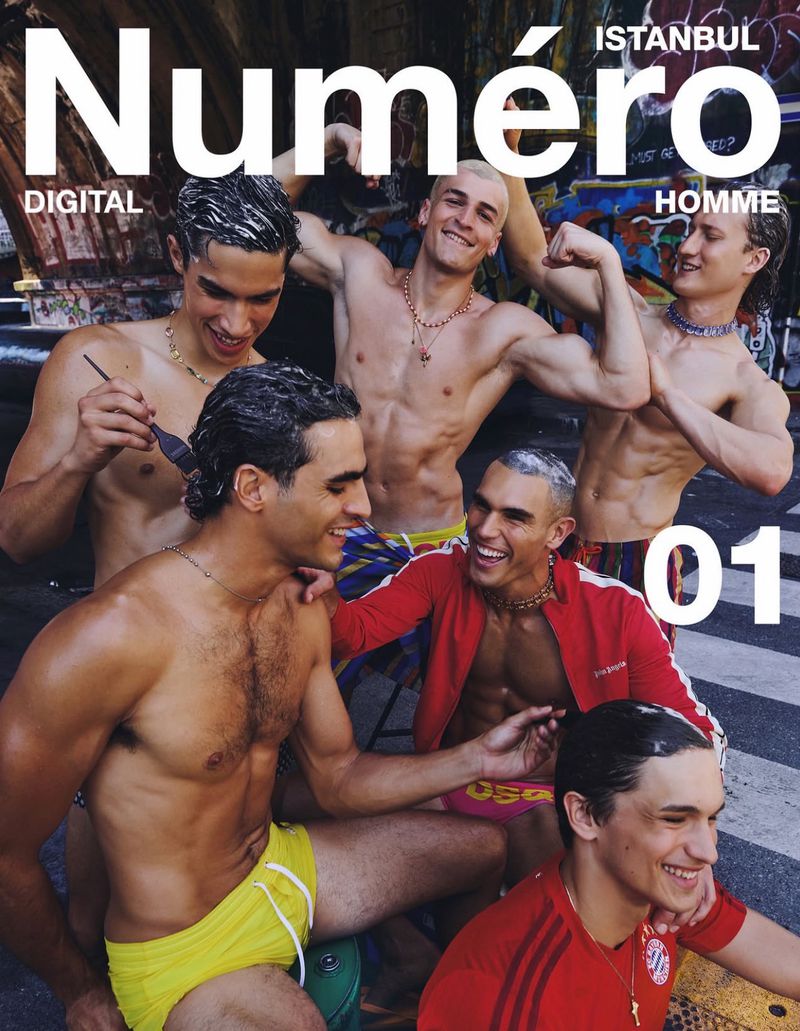Six smiling male models posing together outdoors on a street in colourful swimwear and sportswear for a magazine cover.