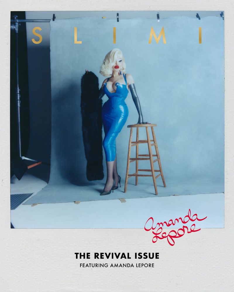 Amanda Lepore in a sculpted blue latex dress standing beside a wooden stool against a studio backdrop on a SLIMI Magazine cover.