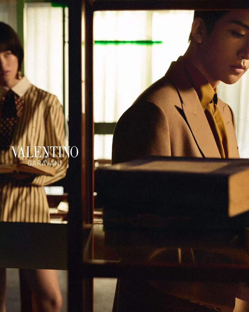 Two models posed in an interior space with bookshelves, featuring Valentino branding overlay