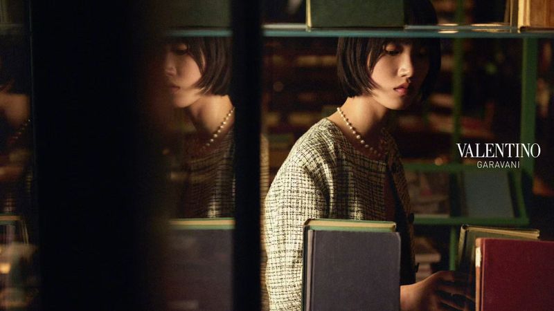 Model Yidan Huang wearing a tweed jacket and pearl necklace looking at books between dark library shelves for Valentino.