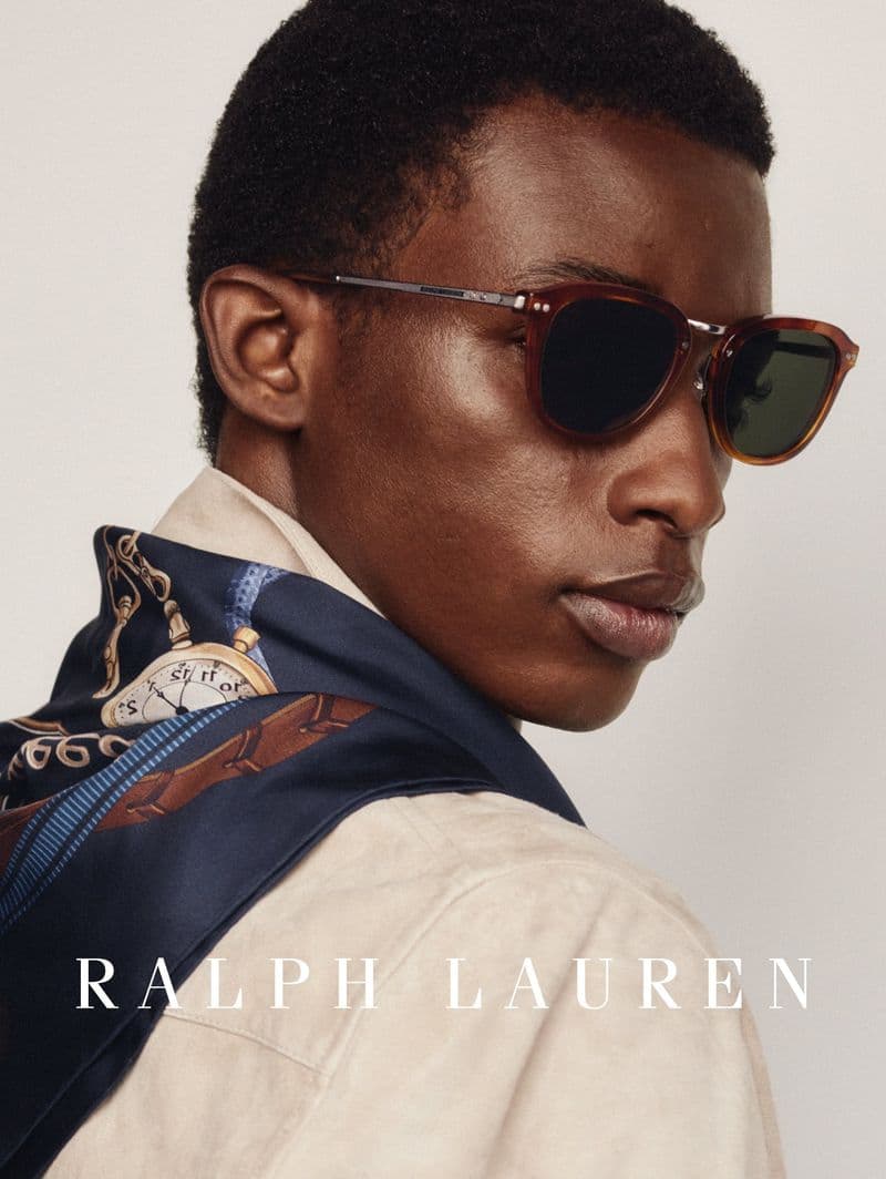 Close-up portrait of a Black man wearing dark sunglasses, a printed silk scarf, and a light beige jacket against a seamless background.