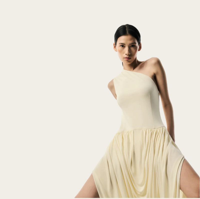 Model Jiayun Xie in a cream one-shoulder draped dress posing against an off-white studio background for Brandon Maxwell.